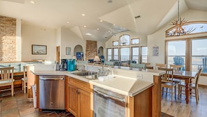 A spacious kitchen with a large island, wooden cabinets, and a dishwasher. There are multiple windows providing natural light and a view of the outdoors. - The Lodge | Oceanfront | Private Pool, Hot Tub | Nags Head (Nags Head)