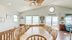 A dining area with a long wooden table and chairs, a ceiling fan, and a view of the ocean through large windows. - Sandpiper's Paradise | Oceanfront | Hot Tub | Kill Devil Hills (Kill Devil Hills)