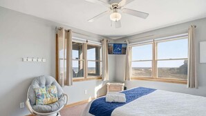 A bedroom with a bed, a chair, a ceiling fan, and large windows. - Sandpiper's Paradise | Oceanfront | Hot Tub | Kill Devil Hills (Kill Devil Hills)