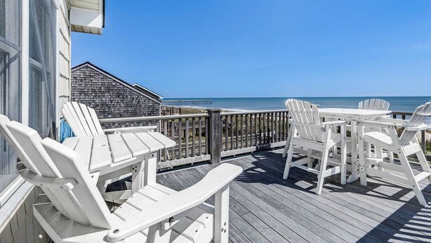 A wooden deck with white Adirondack chairs and a table, overlooking a beach and ocean. - Sandpiper's Paradise | Oceanfront | Hot Tub | Kill Devil Hills (Kill Devil Hills)