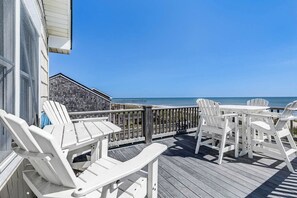A wooden deck with white Adirondack chairs and a table, overlooking a beach and ocean. - Sandpiper's Paradise | Oceanfront | Hot Tub | Kill Devil Hills (Kill Devil Hills)