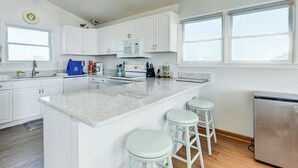A modern kitchen with white cabinets, a central island with stools, a microwave, and a stainless steel refrigerator. - Sandpiper's Paradise | Oceanfront | Hot Tub | Kill Devil Hills (Kill Devil Hills)