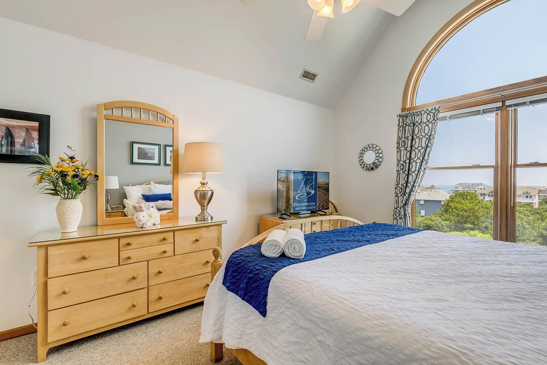 A bedroom with a large bed, a wooden dresser, a mirror, a TV, and a view of the city through a large window.