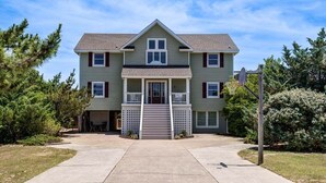 A two-story residential building with a front porch, a garage, and a basketball hoop. - Legacy | 980 ft from the beach | Dog Friendly, Private Pool, Hot Tub | Corolla (Corolla)