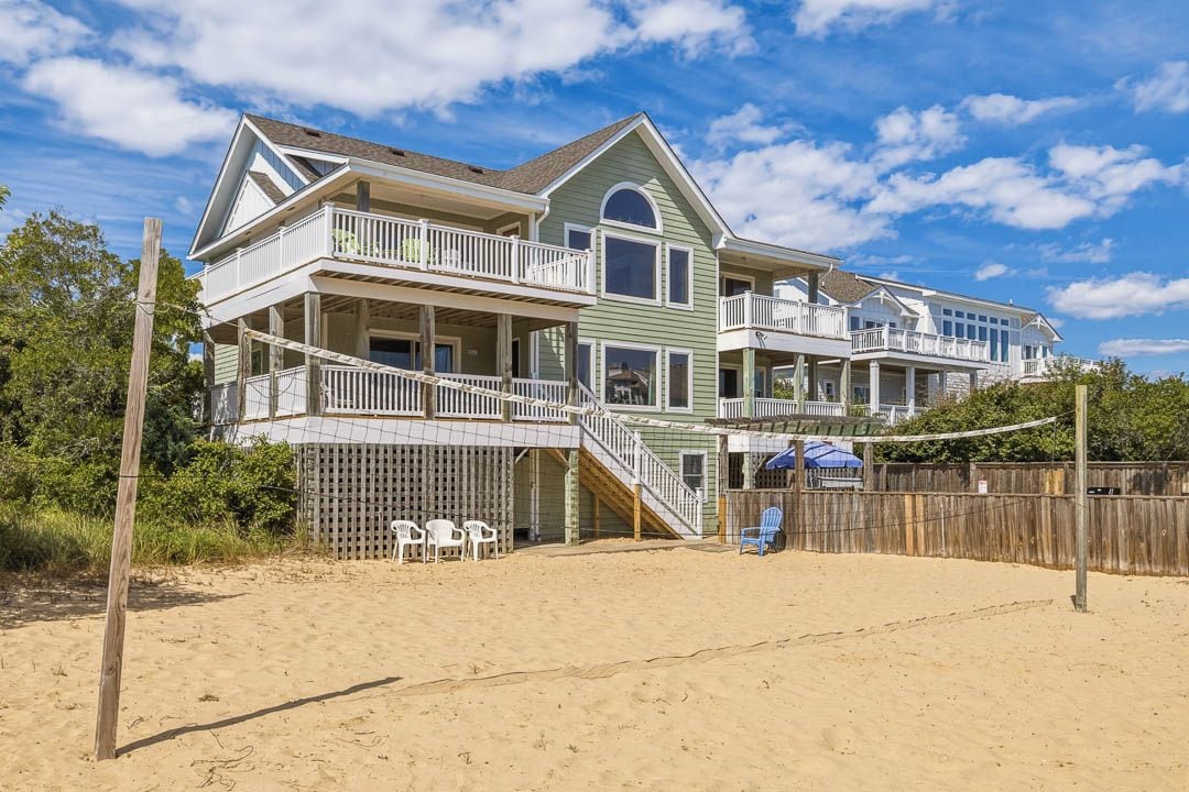 A large two-story beachfront house with multiple balconies, a wooden fence, and beach chairs.