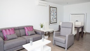 Una sala de estar moderna con un sofá gris, una mesita de centro blanca y un sillón gris. En la pared, hay una obra de arte abstracta enmarcada y una pequeña planta en maceta sobre la mesita.