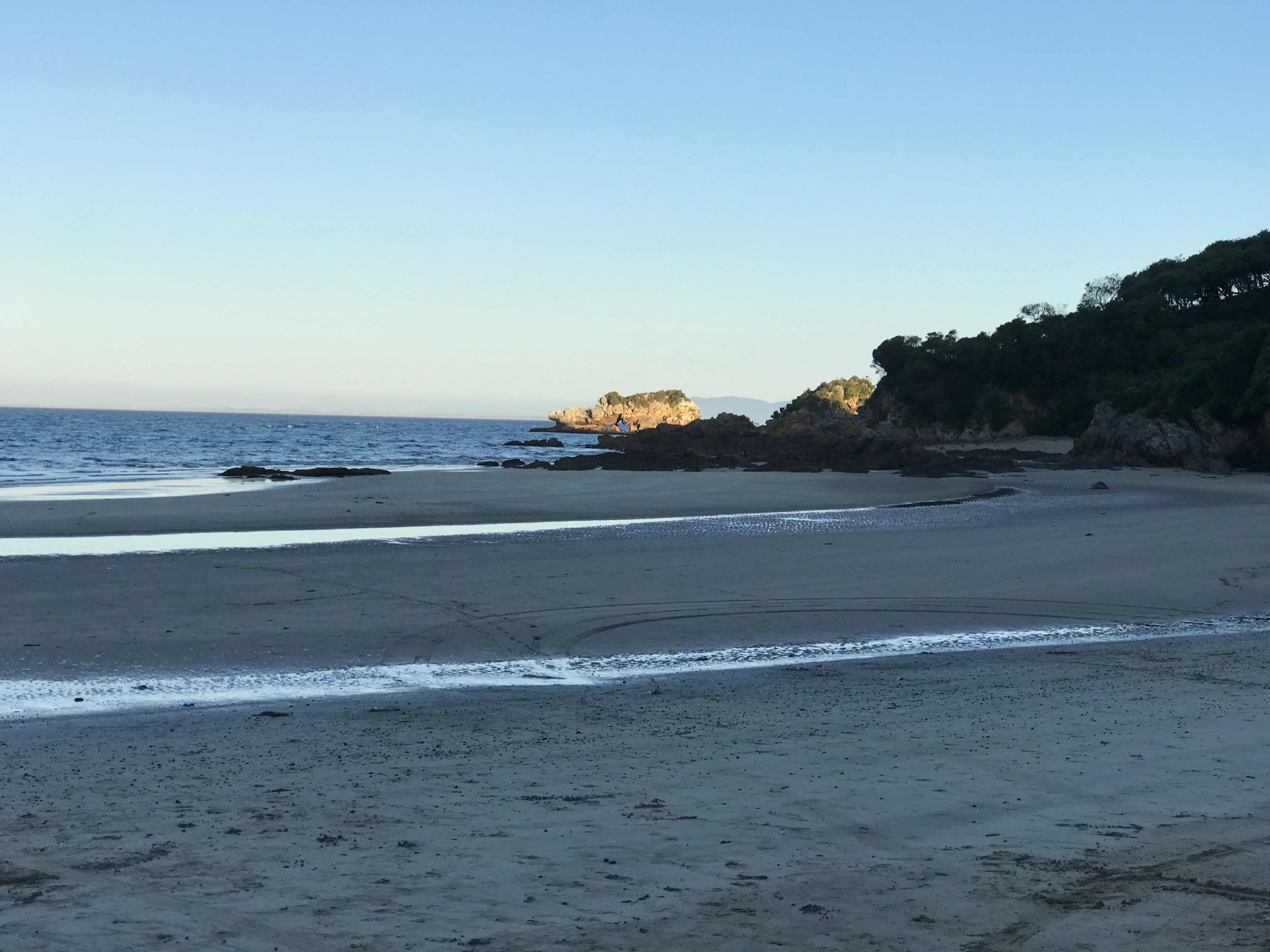 A beach with a rocky outcrop and trees in the background.