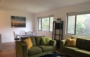 A living room with two green sofas, a dining table with chairs, and large windows with a view of greenery. - Barnacle - a beautiful holiday house in the stunning location of Walkerville (Walkerville)