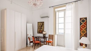 Ein Schlafzimmer mit einem Bett, einem Schreibtisch, zwei Stühlen, einem Fenster mit Vorhängen und einer Wand-Klimaanlage.