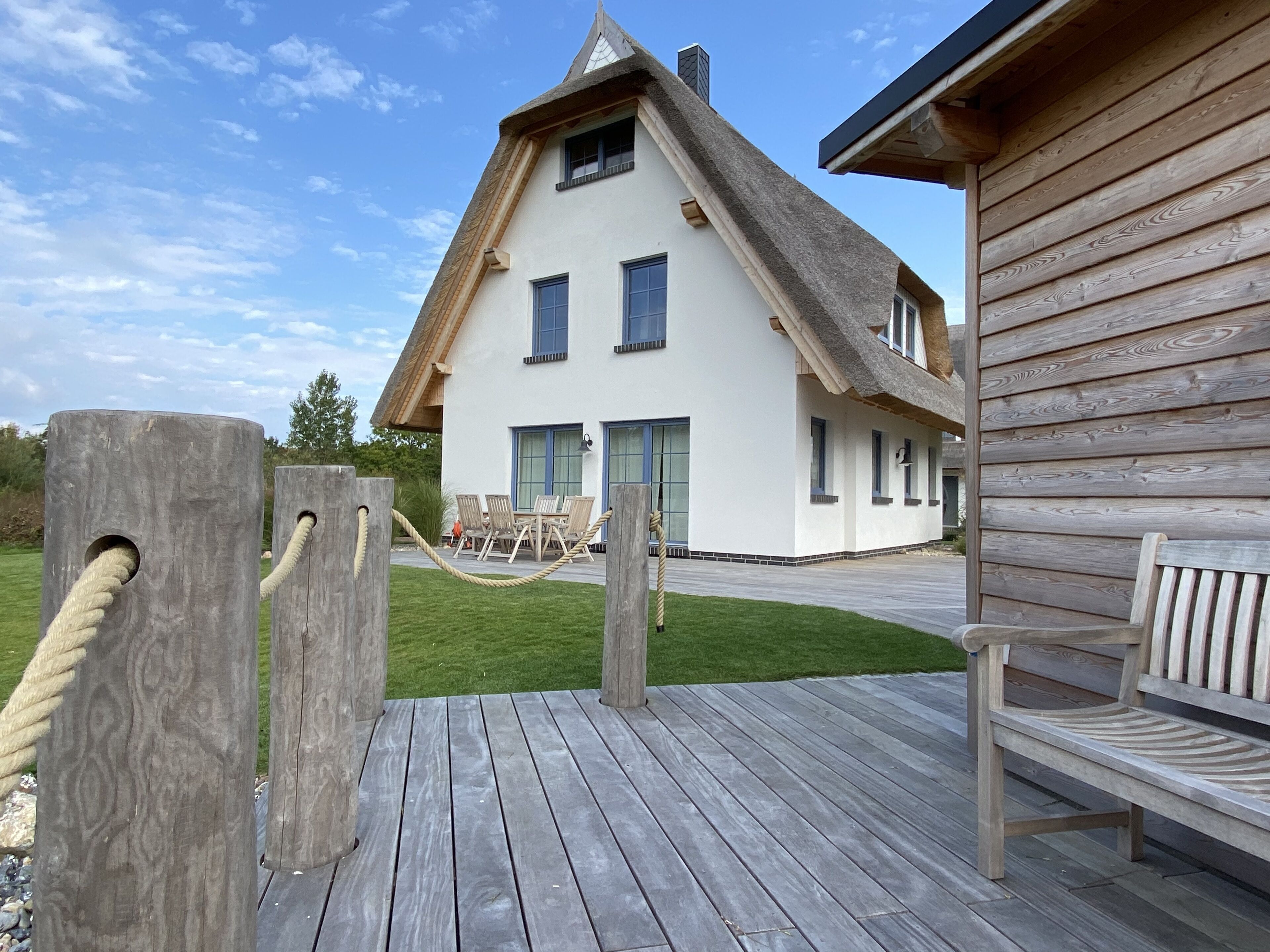 Une terrasse en bois avec un banc, une barrière en corde et une maison blanche dotée d’un toit en pente et de fenêtres.