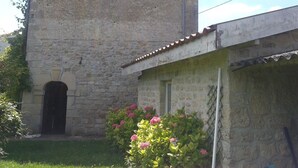 A stone building with a small window and a door, surrounded by a garden with pink flowers.