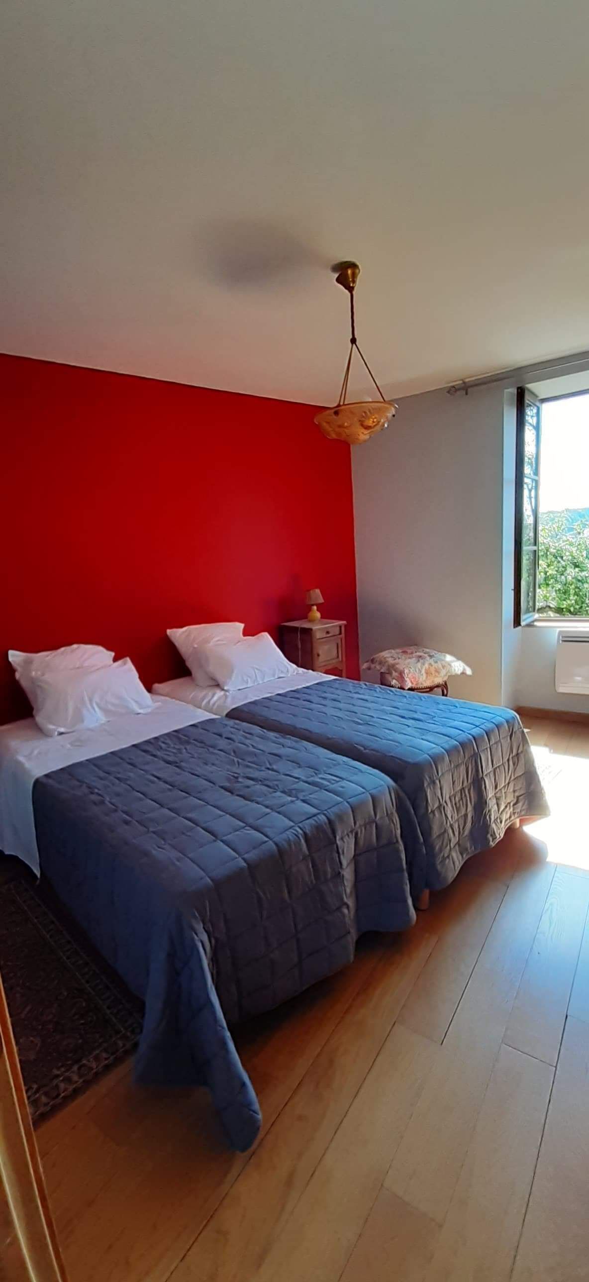 Een slaapkamer met twee bedden, een rode accentmuur, een hanglamp en een raam met uitzicht.