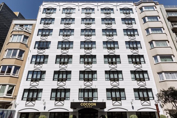 A multi-story hotel with a uniform facade, featuring a series of windows and balconies. The hotel's name, "COCOON," is prominently displayed at the ground level. - Hotel Cocoon (Ostend)