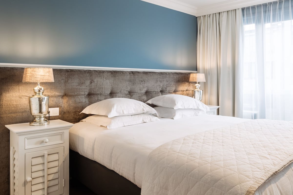 A neatly made bed with a quilted white duvet, a wooden nightstand with a lamp, and a headboard against a blue wall.