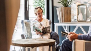 A person reading a magazine in a room with a window, a plant, and a lamp on a table. - Hotel Cocoon (Ostend)