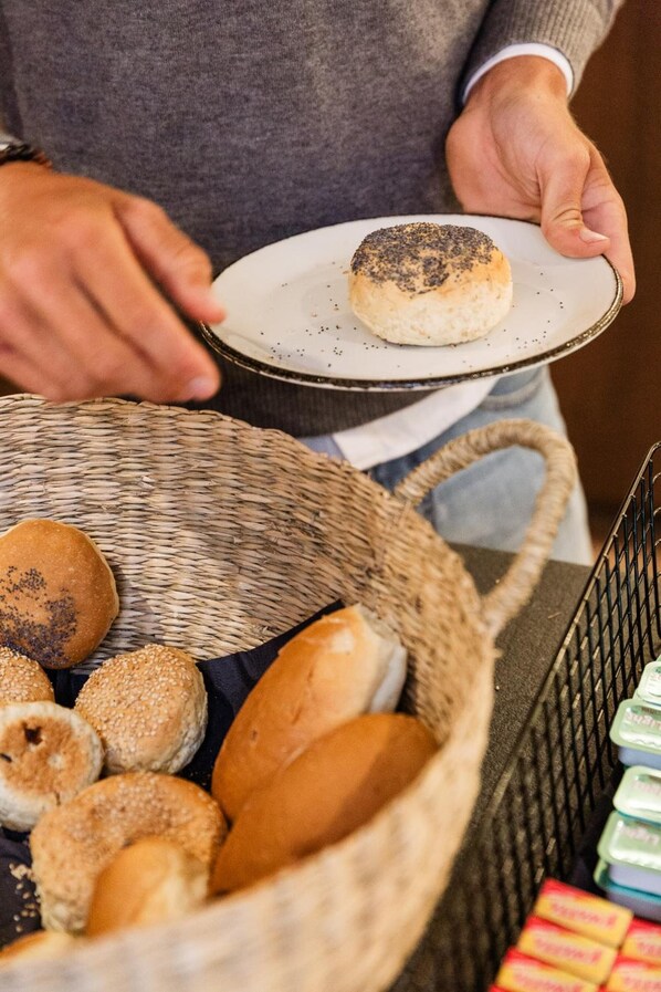 A person holding a plate with a poppy seed bun, with a basket of various buns and a tray of packaged items in the foreground.