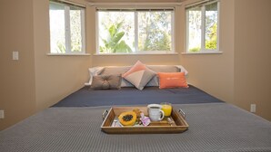 Ein Hotelzimmer mit einem Bett, zwei Fenstern mit Jalousien, einem Tablett mit Frühstücksangeboten und Blick auf Grünflächen.