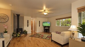 Ein Wohnzimmer mit Kamin, Fernseher, Deckenventilator und Fenster mit Blick auf Grünflächen.