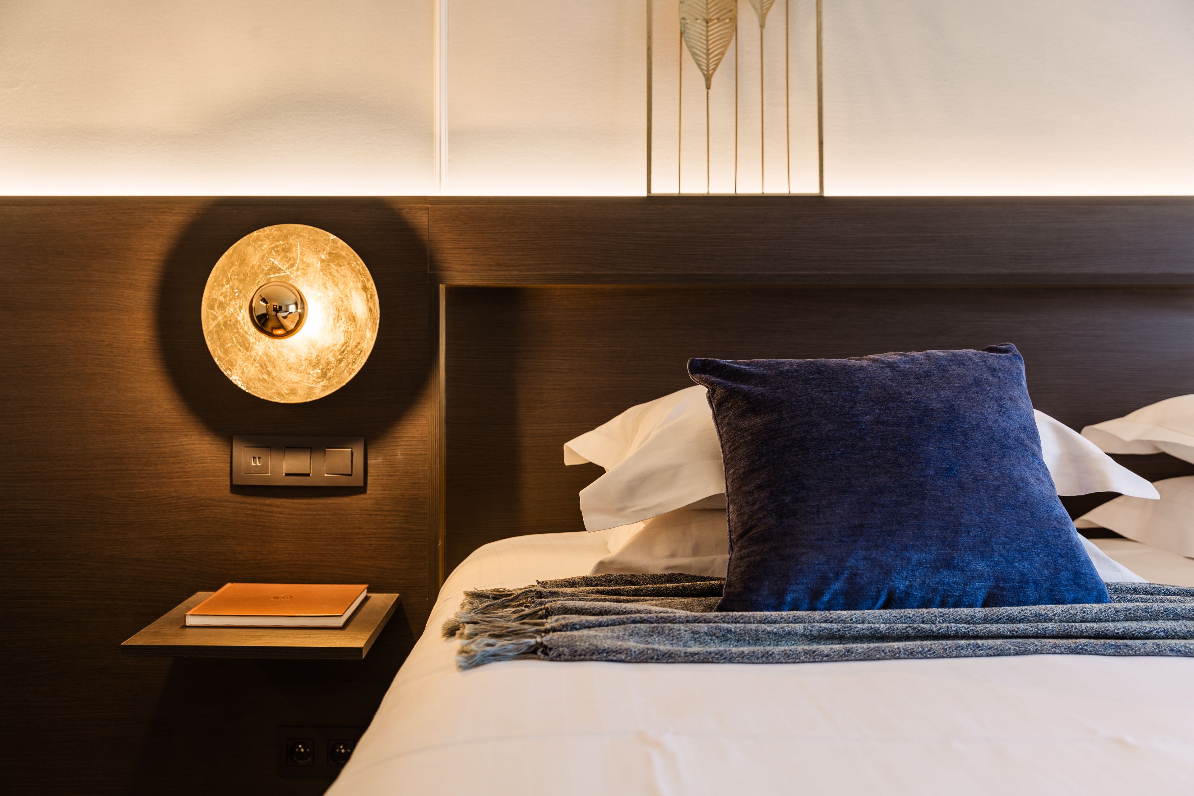 A neatly made bed with a blue pillow and a white pillow, a bedside table with a book, and a wall-mounted light fixture.