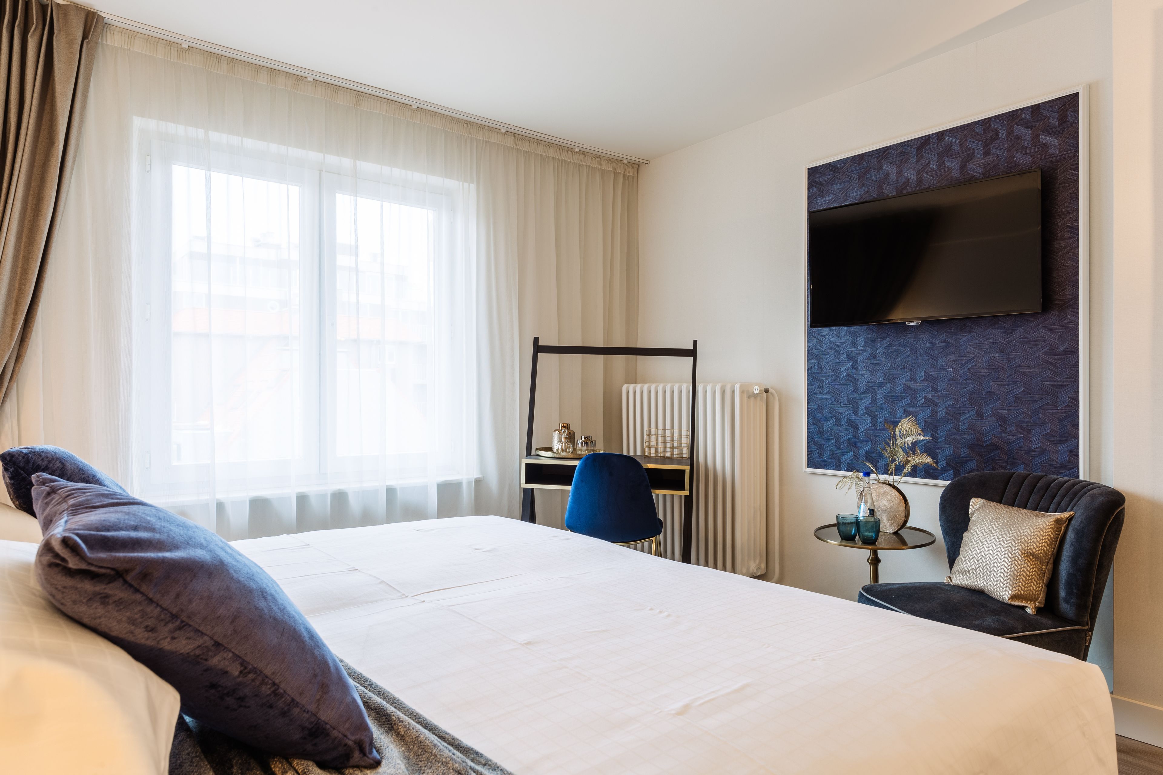 A hotel room with a large bed, a desk, a chair, a TV mounted on the wall, and a window with curtains.