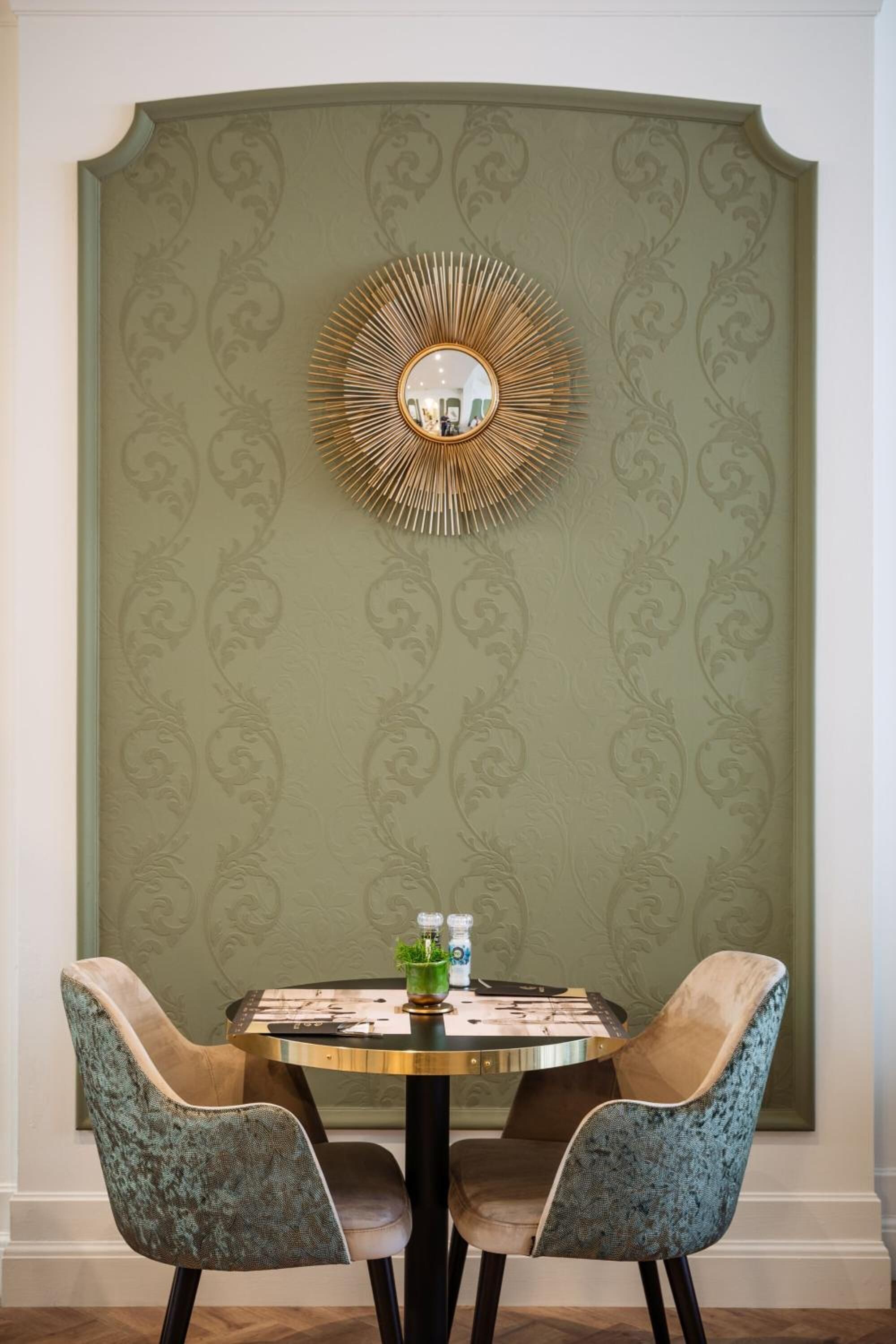 A dining area with a round table, two upholstered chairs, and a decorative mirror on the wall.