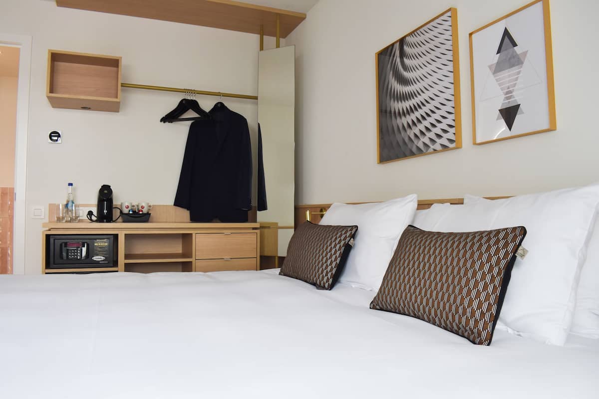 Habitación de hotel con cama, almohadas, mesita de noche con cafetera, microondas, perchero y una obra de arte en la pared.
