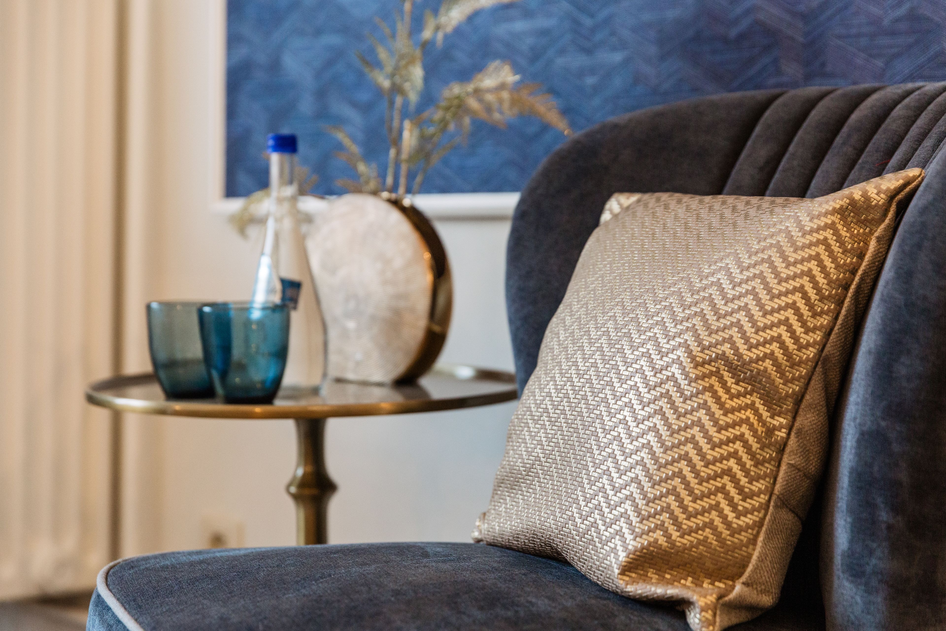 Een donkerblauwe bank met een kussen met goudkleurige textuur, een blauwe glazen vaas, en een witte vaas met gedroogde planten op een klein rond tafeltje.