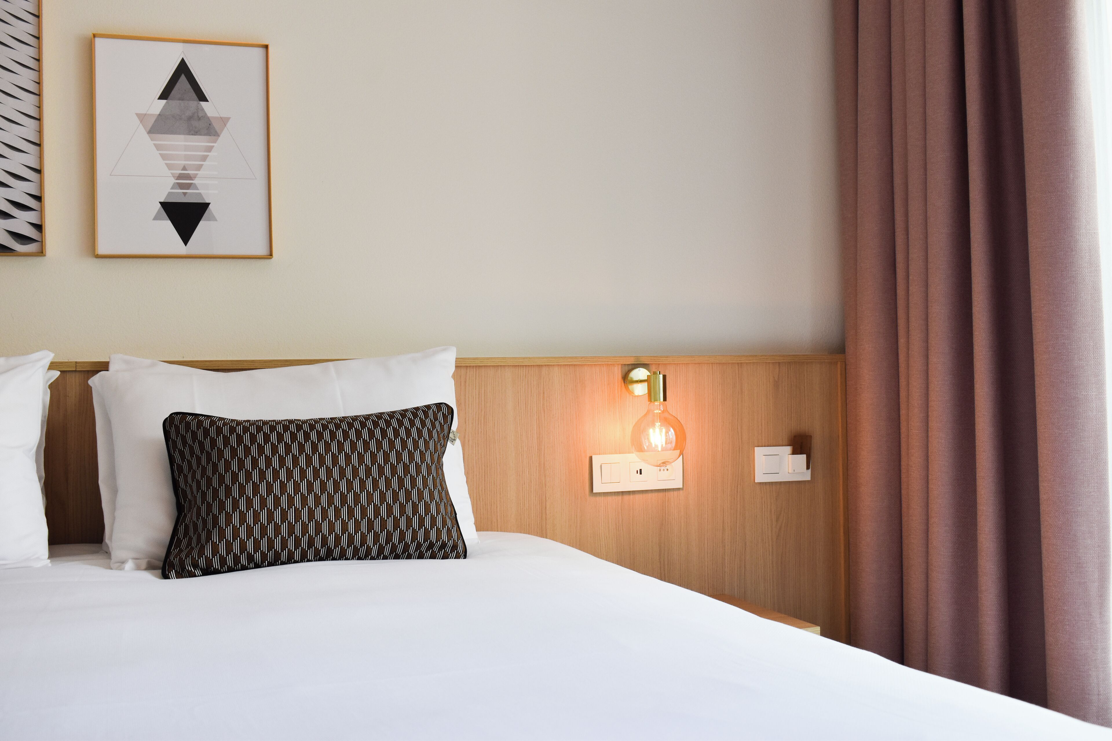 A hotel room with a neatly made bed, a patterned pillow, a wall-mounted light, and a framed geometric artwork.