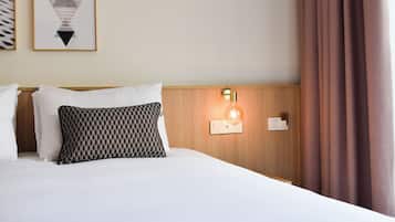A hotel room with a neatly made bed, a patterned pillow, a wall-mounted light, and a framed geometric artwork.