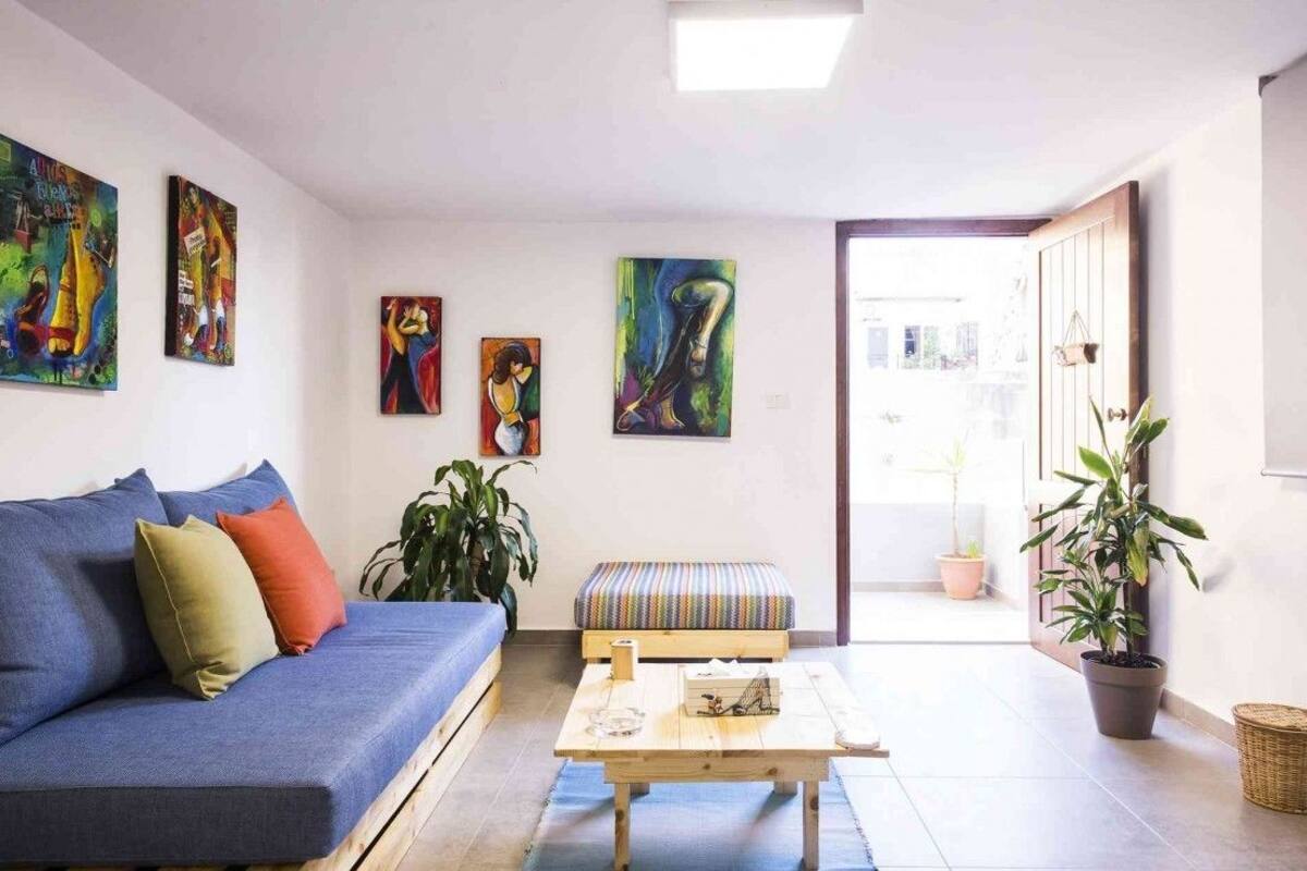 A living room with a blue sofa, a wooden coffee table, and a bench. There are four paintings on the wall and several potted plants.