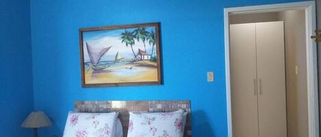 Un dormitorio con una cama, un cuadro de una playa, un aire acondicionado y una puerta a otra habitación.