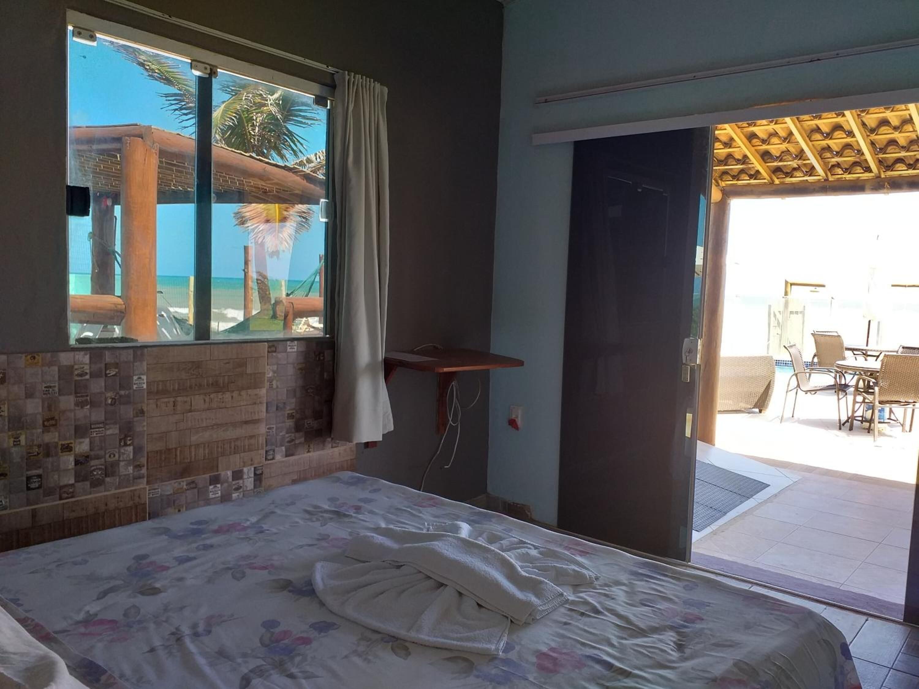 A hotel room with a bed, a window with a view of the beach, a door leading to an outdoor area with chairs and tables.