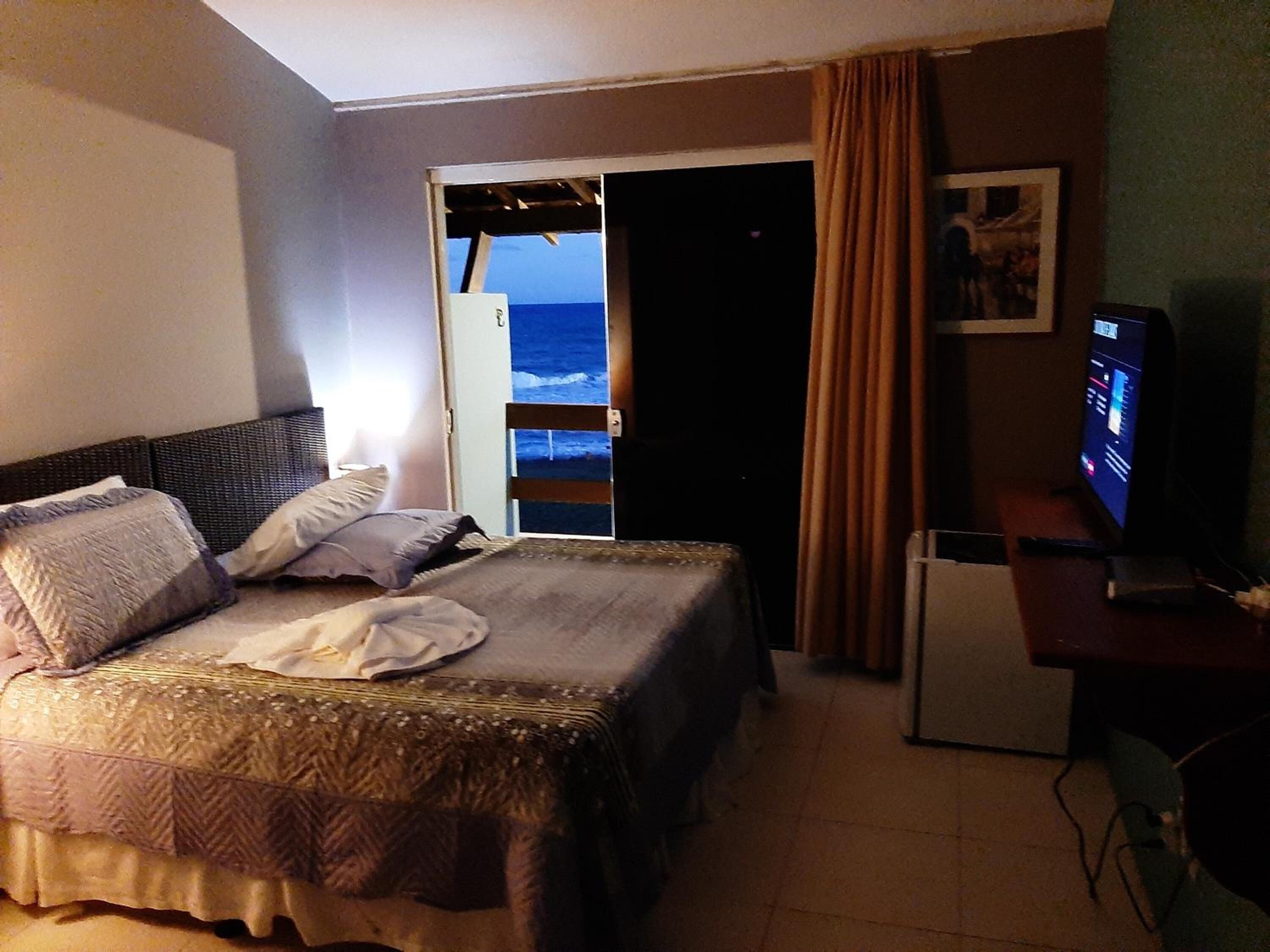 Habitación de hotel con cama, escritorio, televisión y vista al océano a través de una puerta abierta.