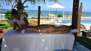 Relax with a soothing massage under a shaded structure overlooking the ocean, surrounded by a tranquil pool area.