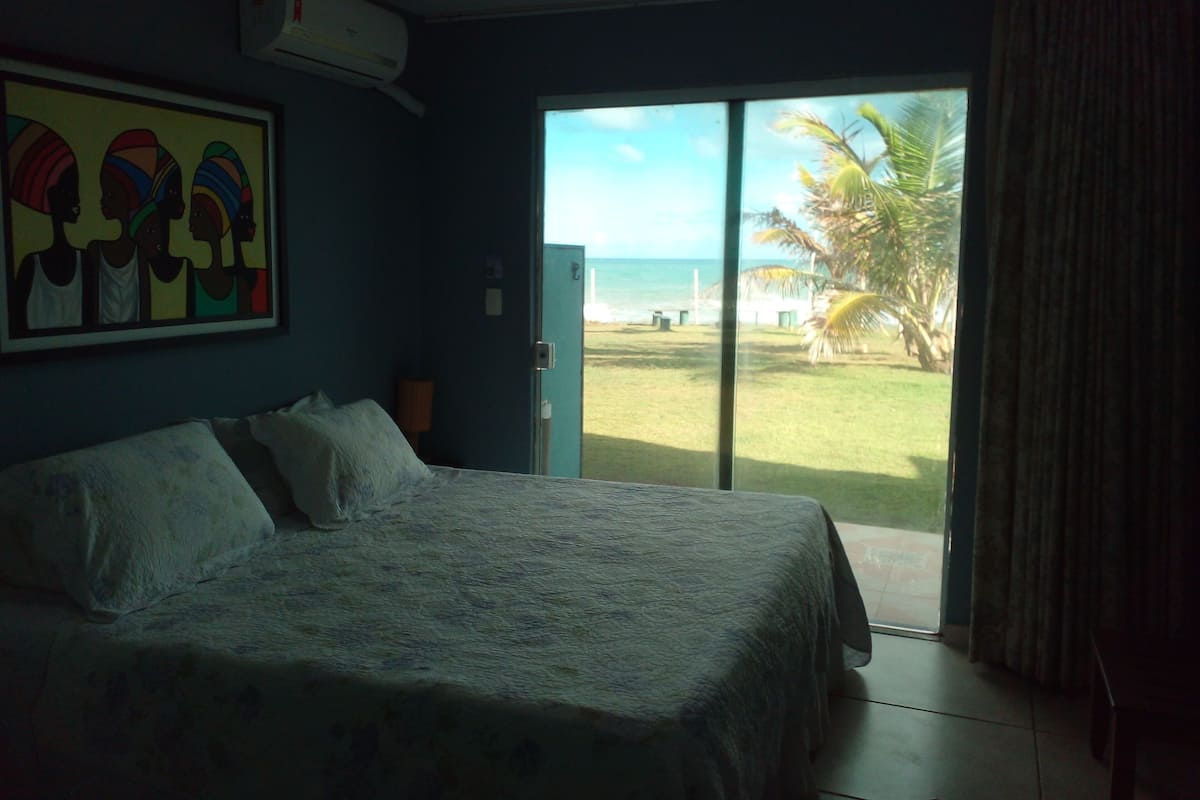 A bedroom with a bed, a painting, and a view of the beach through a sliding door.