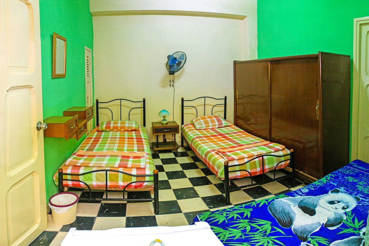 A room with two single beds, a wardrobe, a small table with a lamp, and a fan on the wall.