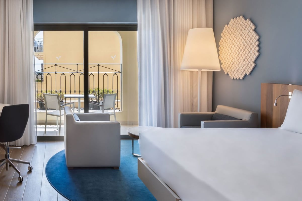 A hotel room with a large bed, a desk, a chair, a sofa, and a balcony with a railing.