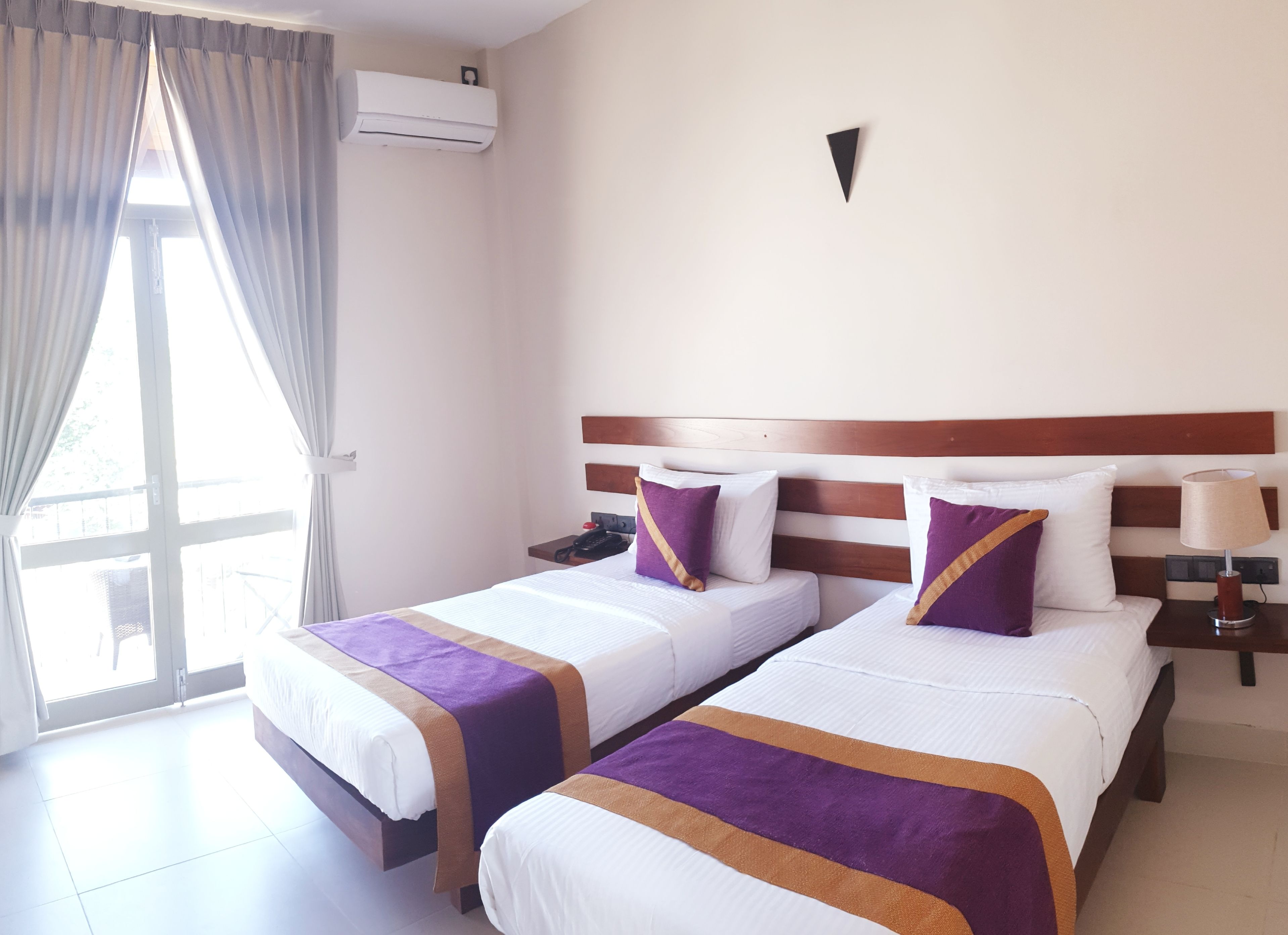 A hotel room with two beds, a wooden headboard, a nightstand with a lamp, and an air conditioning unit.