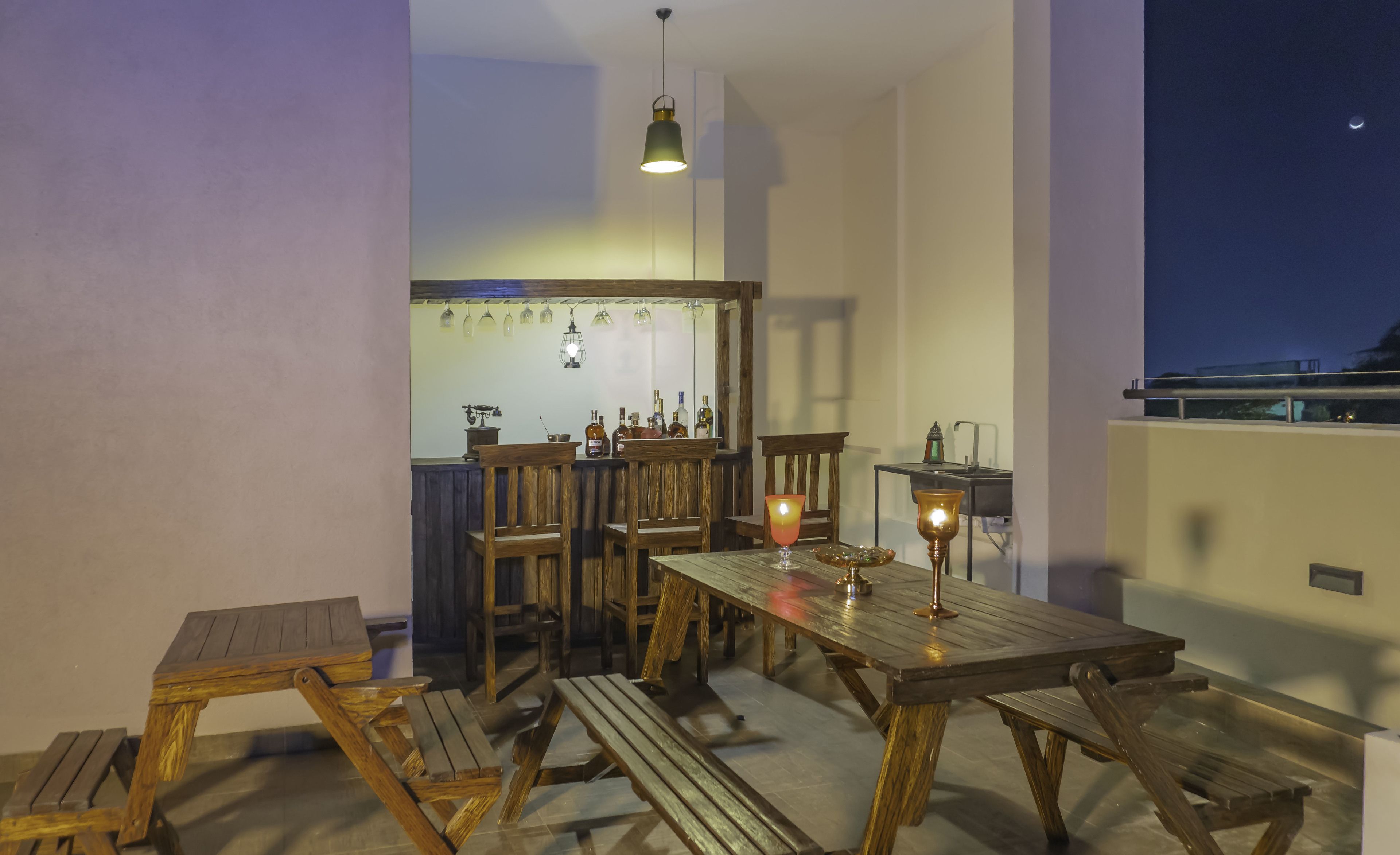 Un espace de restauration extérieur en bois, avec des bancs et une table, un coin bar avec des bouteilles et des verres, et un luminaire suspendu.