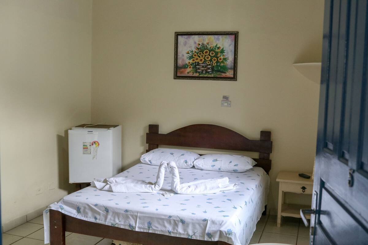 Un lit simple avec une tête de lit en bois, une petite table de chevet, un réfrigérateur blanc et un tableau représentant des fleurs accroché au mur.