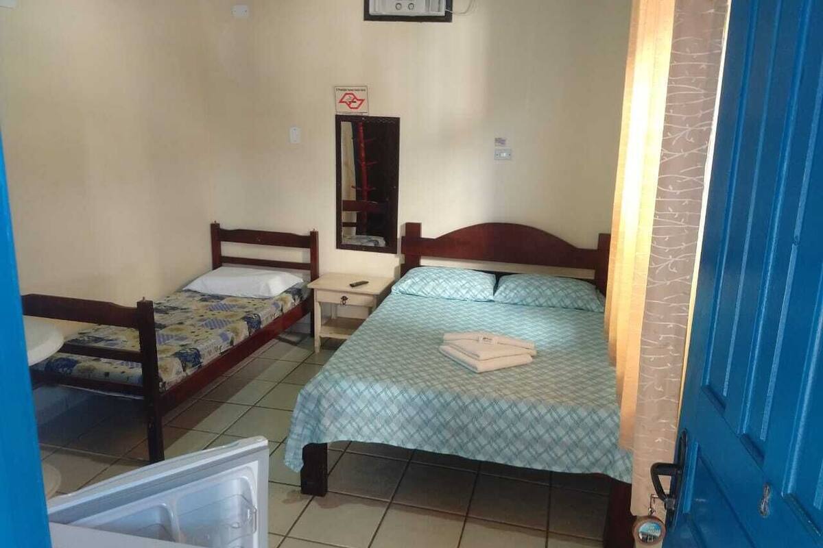 Une petite pièce, de taille modeste, comprenant deux lits simples, une table de chevet, un radiateur fixé au mur et une porte bleue.