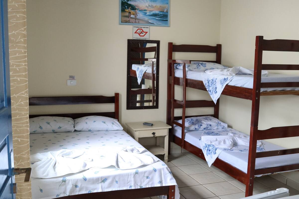 Une chambre avec deux lits, l’un doté d’un cadre en bois et l’autre d’un cadre en métal. On y trouve une petite table avec une chaise, un miroir fixé au mur et un tableau au-dessus des lits.