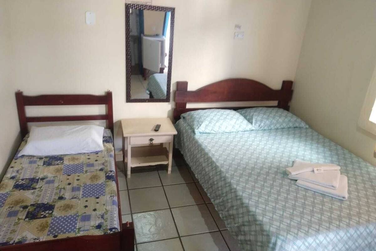 Une petite chambre propre, avec deux lits simples, une table de chevet, un miroir et une fenêtre.