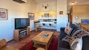 Ein Wohnzimmer mit Fernseher, Couchtisch aus Holz, Ledersofa und einer Küche im Hintergrund.