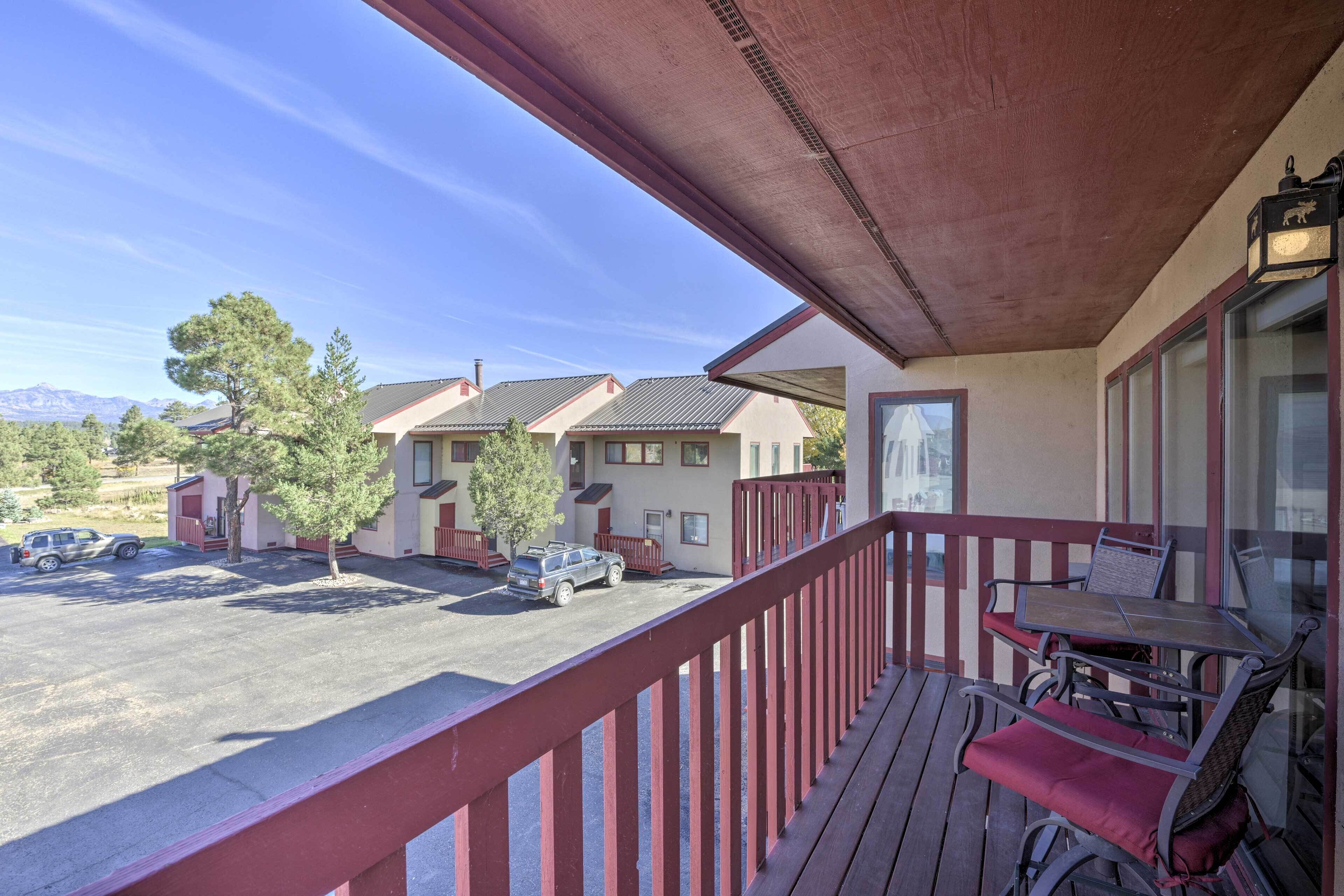 A wooden deck with a table and chairs, overlooking a parking area and buildings with a mountain backdrop.