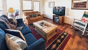 Ein Wohnzimmer mit blauem Sofa, braunem Ledersessel, Couchtisch aus Holz, Fernseher und Schaukelstuhl.
