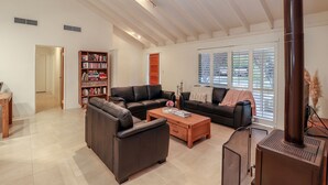 Ein geräumiges Wohnzimmer mit einem großen, schwarzen Ledersofa, einem Holztisch und einem Kamin. Ein Bücherregal voller Bücher und ein Fenster mit Jalousien sind ebenfalls vorhanden.