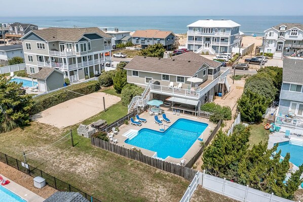 A residential area with multiple two-story houses, a large swimming pool, and a beach in the background. - Joy Seas | 265 ft from the beach | Private Pool (Virginia Beach)