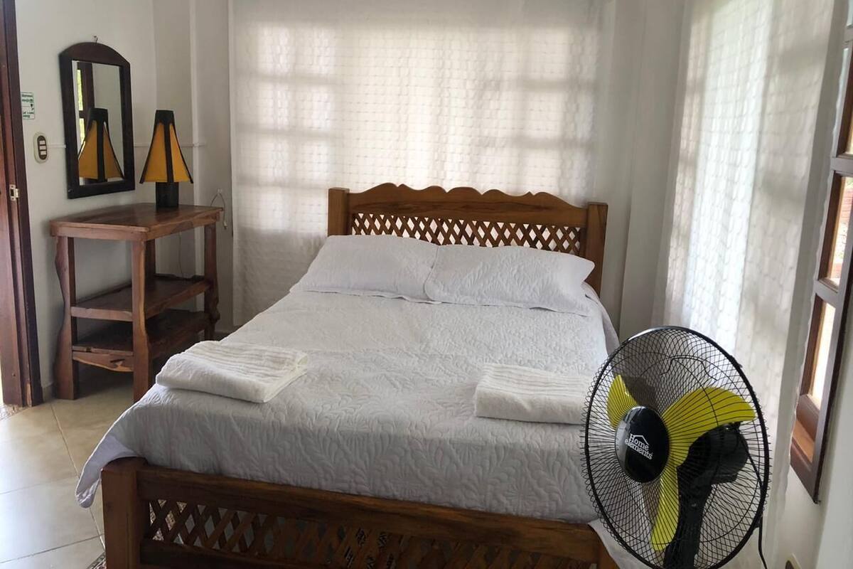 A neatly made bed with a wooden headboard, a bedside table with a lamp, and a standing fan.