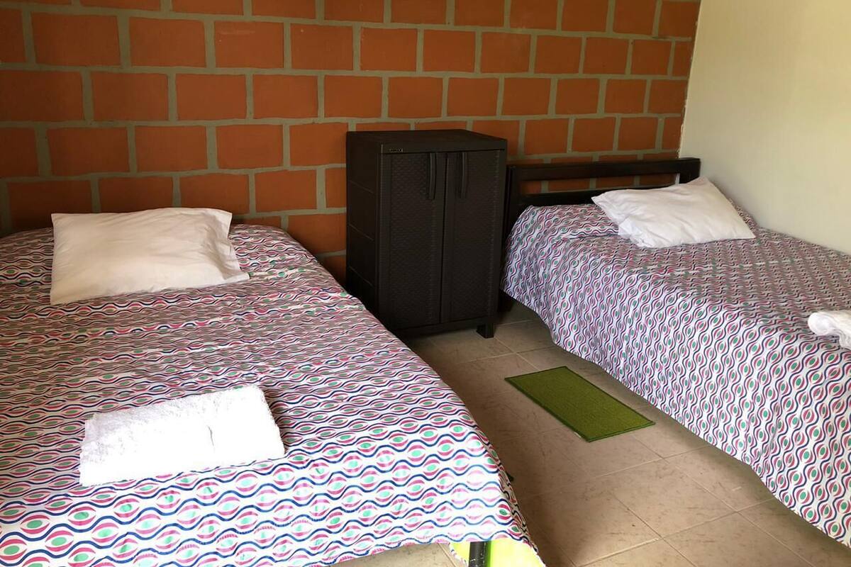A room with two beds, each with patterned bedding and pillows, a black wardrobe, and a green mat on the floor.